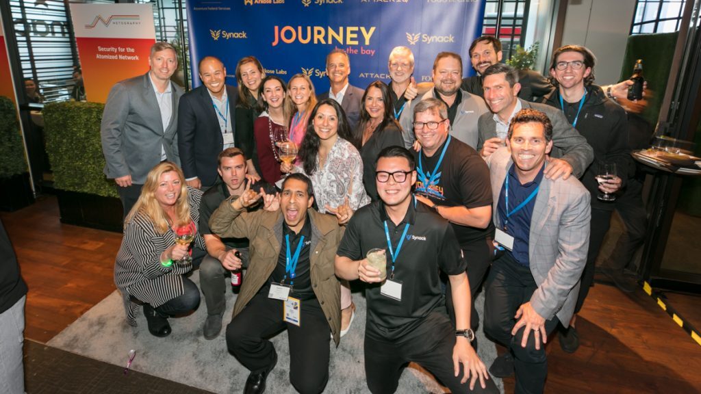 Synack employees grouped together to pose for a photo in front of a step and repeat banner at the Fogo de Chao restaurant in San Francisco.