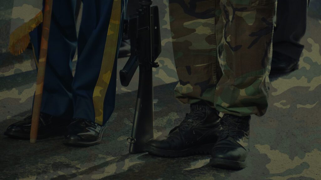 An image of two military members legs with a rifle and flag, overlaid by camo