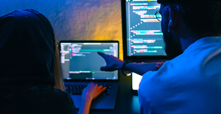 An image of two people sitting at two computers working together in a dark room, illuminated by the light from the computer.